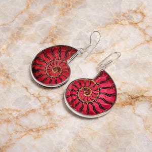 May include: Silver earrings with a red and brown nautilus shell design. The earrings have a spiral pattern with a silver border and a hook closure. The background is a marble-effect surface.