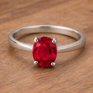 May include: A silver ring with a large, oval-cut, bright red gemstone. The gemstone is held in place by four prongs. The ring is set against a wooden background.