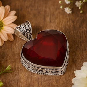 Puede incluir: Colgante en forma de corazón con una gran piedra preciosa roja oscura facetada. El colgante está engastado en metal plateado con detalles ornamentados. Flores sobre una superficie de madera.