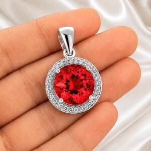 May include: A circular pendant featuring a large, round, vibrant red gemstone set in a polished silver-toned metal frame. The center stone is surrounded by a halo of small, clear, sparkling stones, and the piece includes a silver-toned bail.