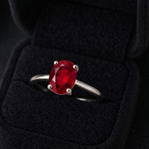 May include: A silver ring with a large, oval-cut ruby gemstone. The vibrant red stone is set with four prongs, and the ring is displayed in a black velvet ring box. The ring is a classic piece of jewelry.