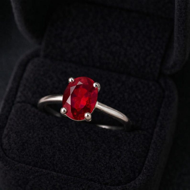 May include: A silver ring with a large, oval-cut ruby gemstone. The vibrant red stone is set with four prongs, and the ring is displayed in a black velvet ring box. The ring is a classic piece of jewelry.