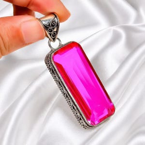 Op de afbeelding: Een rechthoekige hanger met een grote, helder roze edelsteen. De edelsteen is gezet in een zilverkleurig metalen frame met ingewikkelde filigraan details. De hanger wordt door een hand vastgehouden tegen een witte, zijdeachtige achtergrond.