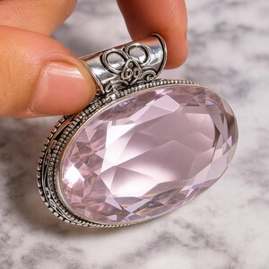 Puede incluir: Un colgante grande, de forma ovalada, con una piedra preciosa facetada de color rosa pálido. La piedra preciosa está engastada en un marco de metal plateado con detalles ornamentados. La fianza presenta un diseño decorativo. El colgante es sostenido por una mano.