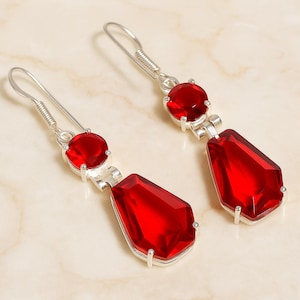 May include: A pair of silver-toned drop earrings featuring vibrant red, faceted gemstones. Each earring has a round gem at the top and a larger, teardrop-shaped gem below, creating a striking contrast.