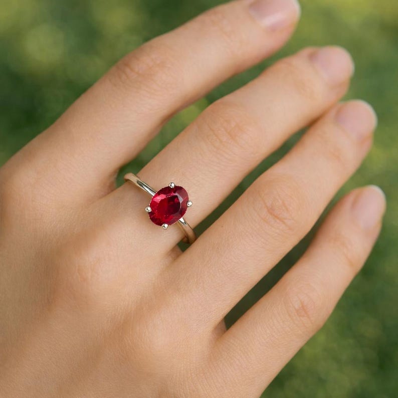 May include: A gold ring featuring a large, oval-cut, deep red gemstone. The gemstone is set with six prongs. The ring is shown on a hand with a blurred green background.