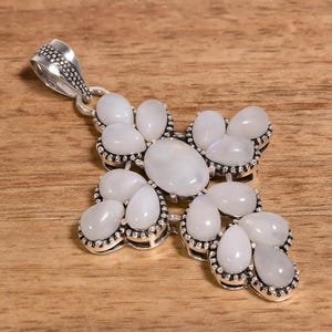 May include: A silver pendant in the shape of a cross, adorned with multiple white, oval-shaped gemstones. The pendant has a textured bail for a chain and is set against a wooden background.