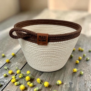 May include: A round, off-white cotton rope basket with a dark brown rim and a matching side handle. A brown faux leather tag is stitched to the rim, featuring the text "MADE WITH" followed by a small heart symbol in black ink.
