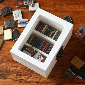 May include: A white miniature bookcase filled with tiny books, with more scattered around on a wooden surface. The books have colorful spines and covers, creating a vibrant display. The bookcase has a hinged door and a latch.