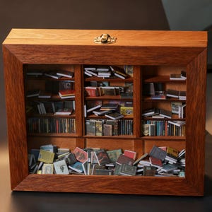 Puede incluir: Una estantería de madera en miniatura con tapa con bisagras y cierre de latón. El interior está lleno de libros diminutos de varios tamaños y colores, dispuestos en estantes y esparcidos en la base. La estantería está hecha de madera teñida.