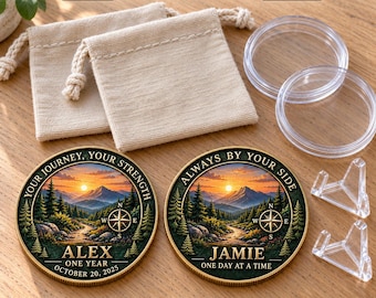 May include: A personalized 2-coin gift set featuring two round coins with mountain landscape imagery, compass icons, and text including names Alex and Jamie. Includes two beige fabric drawstring pouches, two clear plastic capsules, and two clear display stands.