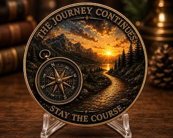 Puede incluir: Un medallón circular en tonos bronce con un paisaje de montañas, un bosque y un camino al atardecer. Una brújula está grabada a la izquierda. El texto dice THE JOURNEY CONTINUES y STAY THE COURSE en letras en relieve.
