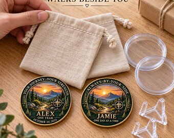 May include: Two round metal coins featuring a colorful mountain landscape with a compass, pine trees, and a sunset. One coin reads "YOUR JOURNEY, YOUR STRENGTH, ALEX, ONE YEAR, OCTOBER 20, 2025". The other reads "ALWAYS BY YOUR SIDE, JAMIE, ONE DAY AT A TIME".