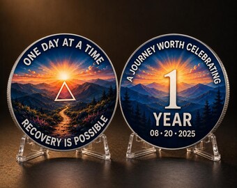 May include: Two circular metal sobriety coins displayed on clear stands. The left coin features a sunrise over mountains, a triangle, and the text ONE DAY AT A TIME RECOVERY IS POSSIBLE. The right coin shows the text A JOURNEY WORTH CELEBRATING 1 YEAR 08 20 2025.