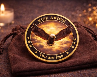 May include: A gold-colored coin with a detailed image of an eagle with outstretched wings, set against a cloudy sky. The coin is inscribed with "RISE ABOVE" and "You are free." The coin rests on a brown drawstring bag.