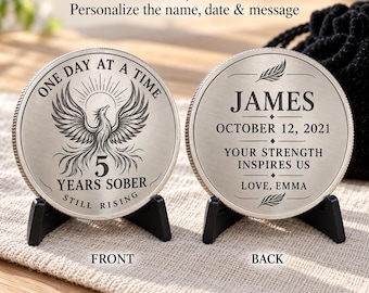 May include: Two silver-toned metal sobriety coins displayed on black stands. The front features a phoenix, the text ONE DAY AT A TIME, 5 YEARS SOBER, and STILL RISING. The back displays JAMES, OCTOBER 12, 2021, YOUR STRENGTH INSPIRES US, and LOVE, EMMA.