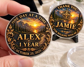 May include: Two round metal commemorative coins featuring a landscape with mountains, a forest, and a sunset. One coin displays the text ONE DAY AT A TIME, ALEX, 1 YEAR, OCTOBER 25, 2025. The other coin displays THANK YOU, JAMIE, FOR WALKING WITH ME.