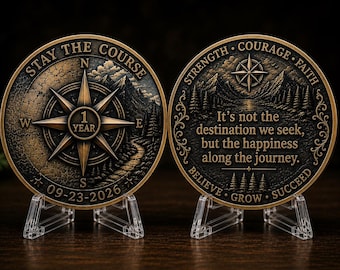 May include: Two circular bronze-toned commemorative coins displayed on clear plastic stands. The left coin features a compass rose, the text STAY THE COURSE, 1 YEAR, and 09-23-2026. The right coin displays a mountain landscape and the text It's not the destination we seek, but the happiness along the journey, STRENGTH, COURAGE, FAITH, BELIEVE, GROW, SUCCEED.