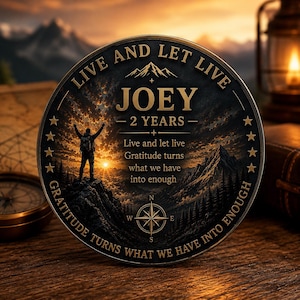 Könnte beinhalten: Eine runde Medaille mit einer Berglandschaft bei Sonnenuntergang und der Silhouette eines Wanderers. Der Text lautet: LIVE AND LET LIVE, JOEY, 2 YEARS und Gratitude turns what we have into enough. Unten befindet sich eine Kompassrose mit Sternen.