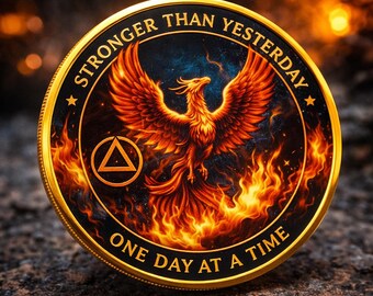 May include: A round, gold-colored coin featuring a phoenix rising from flames. The phoenix is depicted in shades of orange and red against a dark blue and black background. The coin has the text "STRONGER THAN YESTERDAY" and "ONE DAY AT A TIME".
