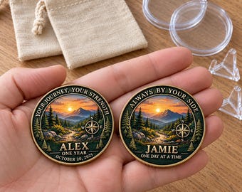 May include: Two round metal coins featuring a landscape illustration of mountains, pine trees, and a compass. One coin reads "YOUR JOURNEY, YOUR STRENGTH, ALEX, ONE YEAR, OCTOBER 20, 2025". The other reads "ALWAYS BY YOUR SIDE, JAMIE, ONE DAY AT A TIME".