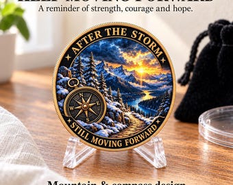 May include: A round commemorative coin featuring a mountain landscape with snow, pine trees, and a golden compass. The text reads AFTER THE STORM and STILL MOVING FORWARD. The coin is displayed on a clear plastic stand with a gold-toned rim.