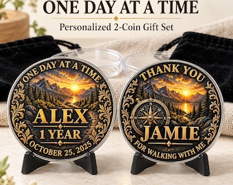 May include: Two round metal coins in black stands, featuring a sunset mountain landscape. The left coin reads "ONE DAY AT A TIME", "ALEX", "1 YEAR", and "OCTOBER 25, 2025". The right coin reads "THANK YOU", "JAMIE", and "FOR WALKING WITH ME".
