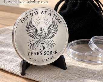 May include: A circular silver-toned sobriety coin featuring a laser-engraved phoenix rising above the number 5. The text reads ONE DAY AT A TIME, 5 YEARS SOBER, and STILL RISING. The coin is displayed on a black stand with a black pouch.
