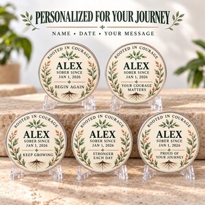 Può includere: Cinque gettoni circolari su supporti trasparenti, ognuno con sfondo crema, bordo di foglie verdi e il testo "ROOTED IN COURAGE", "ALEX", "SOBER SINCE JAN 1, 2026" e frasi come "BEGIN AGAIN".