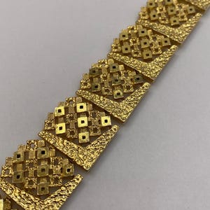Könnte beinhalten: Ein goldfarbenes Armband mit geometrischem Design. Das Armband besteht aus rautenförmigen Gliedern mit strukturierter Oberfläche und quadratischen Aussparungen. Die Glieder sind durch kleinere, rechteckige Teile verbunden.