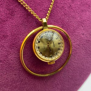 Könnte beinhalten: Eine goldfarbene Anhängeruhr mit rundem Zifferblatt und goldener Kette. Das Zifferblatt hat einen goldenen Hintergrund mit schwarzen Stundenmarkierungen und Zeigern. Die Uhr ist in einem goldenen kreisförmigen Rahmen aufgehängt.