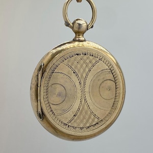 May include: A round gold-toned metal pocket watch case featuring an intricate engraved pattern on the front cover. The design includes two circular motifs with fine concentric lines and a cross-hatched grid section, topped with a circular bail.