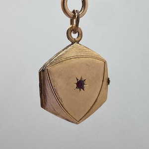 Könnte beinhalten: Ein goldfarbenes, aufklappbares Medaillon mit einem sternförmigen, roten Edelstein in der Mitte. Das Medaillon hängt an einer goldenen Kette mit einem Verschluss. Das Medaillon hat eine geometrische Form mit einem geriffelten Design an den Seiten.