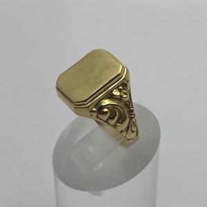 Puede incluir: Un anillo de sello dorado con una cara rectangular y adornos de volutas en los laterales. El anillo se exhibe sobre un soporte cilíndrico transparente. La cara del anillo mide aproximadamente 1,3 cm por 1 cm.