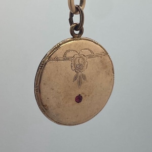 Op de afbeelding: Een goudkleurige, ronde hanger met een bloemmotief en een kleine, rode edelsteen. De hanger hangt aan een ketting. Het bloemmotief is gecentreerd op de hanger en is omgeven door een geëtste rand.