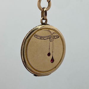 Könnte beinhalten: Ein goldfarbenes, rundes Medaillon mit einem dekorativen Schleifendesign und zwei herabhängenden roten Edelsteinen. Das Medaillon ist an einer goldenen Kette mit einem Verschluss befestigt. Die Oberfläche des Medaillons ist glatt, mit einem detaillierten Rand.