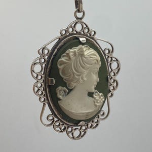 Könnte beinhalten: Ovaler Cameo-Anhänger mit weißem Frauenprofil auf dunkelgrünem Hintergrund, gefasst in einem silberfarbenen Filigranrahmen. Der Anhänger hängt an einer goldfarbenen Kette.