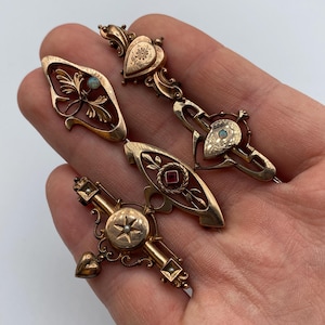 Könnte beinhalten: Vier antike goldfarbene Broschen mit aufwendigen Designs. Die Broschen weisen Herz-, Blatt- und geometrische Formen auf, einige mit kleinen Edelsteinen. Die Broschen werden auf einer Hand präsentiert.