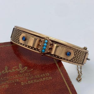 Puede incluir: Un brazalete rígido articulado de tono dorado con un patrón de tejido de cesta texturizado, dos piedras azules pequeñas y una barra central vertical engastada con cuatro cuentas diminutas de color turquesa. Incluye una cadena de seguridad.