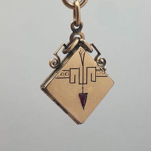 Könnte beinhalten: Ein rautenförmiger Goldmedaillon-Anhänger mit einer zentralen, dunkelroten, dreieckigen Edelsteineinlage. Die Oberfläche ist mit schwarzen, gravierten geometrischen Linien und kleinen kreisförmigen Akzenten verziert. Das Medaillon hängt an einem goldenen Ring.