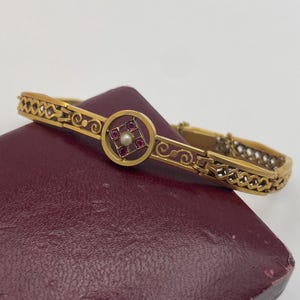 Könnte beinhalten: Ein goldfarbenes Armband mit einem zentralen kreisförmigen Design mit einer Perle und roten Edelsteinen. Das Armband hat aufwendige Filigran- und geometrische Muster. Der Verschluss ist sichtbar, und das Armband liegt auf einer weinroten Oberfläche.