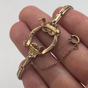 Könnte beinhalten: Ein goldfarbenes antikes Uhrenarmband mit Hufeisen-Design. Das Armband verfügt über einen Klappverschluss, eine kleine Kette und einen kreisförmigen Aufsatz. Die aufwendigen Details und das gealterte Aussehen deuten auf ein Vintage-Stück hin.