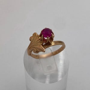 Könnte beinhalten: Ein goldener Ring mit einem runden, facettierten, dunkelrosa Edelstein in einer Fassung mit sechs Krappen. Die Ringschiene ist mit einem strukturierten, blattförmigen Design verziert, das von einer Seite der Fassung ausgeht und ein detailliertes botanisches Aussehen erzeugt.
