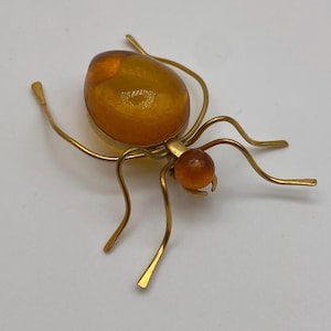 Op de afbeelding: Vintage spinnenbroche. De broche heeft een goudkleurig metalen frame met een amberkleurige, ovale steen voor het lichaam en een kleinere, ronde amberkleurige steen voor de kop. De poten zijn dunne, gebogen gouden draden.
