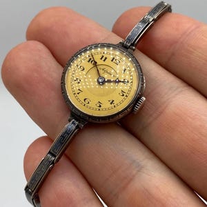 Könnte beinhalten: Eine Vintage-Armbanduhr von Alpina mit einem runden, blassgelben Zifferblatt, schwarzen arabischen Ziffern und dunklen Zeigern. Das Gehäuse und das Gliederarmband aus Metall haben ein dunkles, angelaufenes Finish mit einem geometrischen Mäander-Muster.