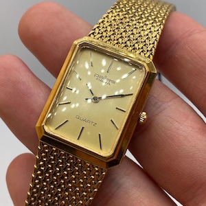 May include: A gold-toned rectangular quartz watch with a mesh band. The watch face is a light cream color with black hands and markers. The watch face reads "pallas paha" and "QUARTZ".