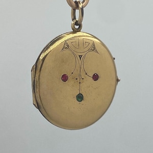 May include: A round gold-toned metal locket featuring an engraved geometric design at the top and three small inset stones, two red and one green, arranged in a triangular pattern on the front surface, suspended from a matching gold-toned metal loop.