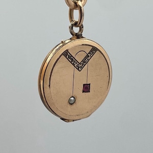 Könnte beinhalten: Ein goldfarbener, runder Anhänger mit dekorativem Design. Der Anhänger hat einen kleinen, quadratischen roten Edelstein und eine perlenartige Perle. Die Oberseite des Anhängers hat ein dekoratives Design. Der Anhänger hängt an einer Goldkette.