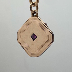 Könnte beinhalten: Ein goldfarbener, geometrischer Anhänger mit einem kleinen, quadratischen, lila Edelstein in der Mitte. Der Anhänger ist an einer Goldkette befestigt. Der Anhänger hat eine rautenförmige Form mit geätzten Linien.