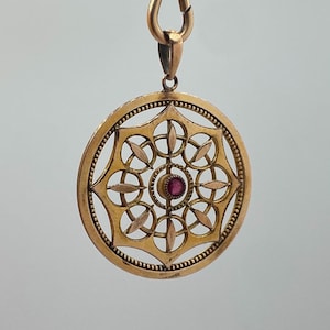 Könnte beinhalten: Ein goldfarbener Anhänger mit einem kreisförmigen, durchbrochenen Design. Er hat einen zentralen, runden, roten Edelstein und ist an einem passenden Verschluss aufgehängt. Das komplizierte Muster umfasst florale und geometrische Formen.